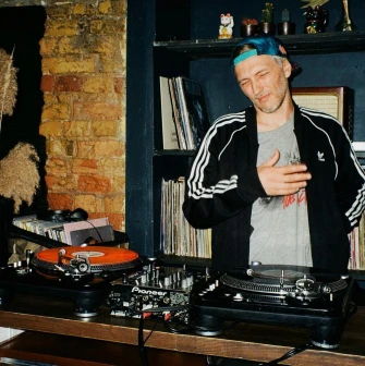 Bar Restaurant Kosatka - man posing in front of two turntables and a shelf with records