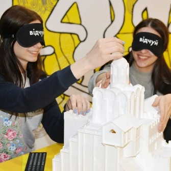 Muzei u Temriavi in Kyiv - smiling visitors wearing black eye masks exploring a 3D white architectural model of a church by touch