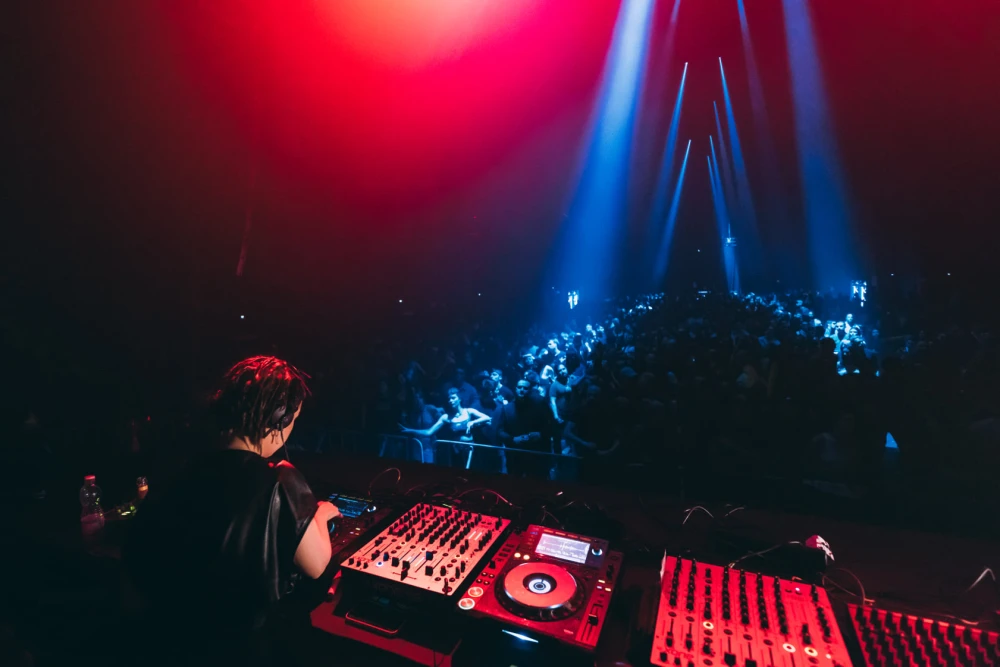 DJ performing at a deck in front of a large crowd with blue light beams