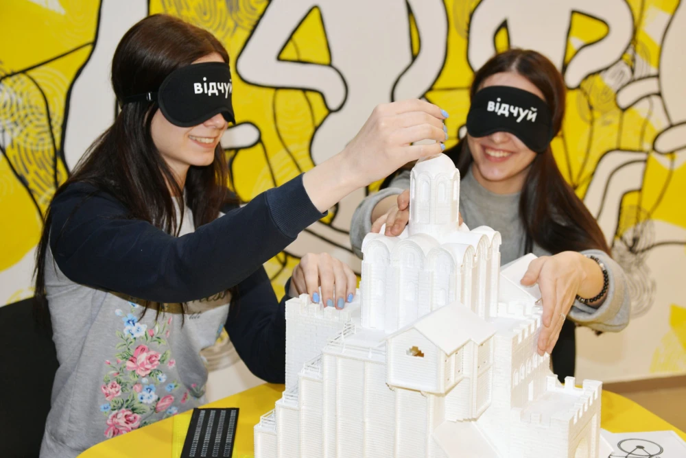 Muzei u Temriavi in Kyiv - smiling visitors wearing black eye masks exploring a 3D white architectural model of a church by touch