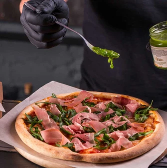 Chef in black gloves adds green pesto sauce to a fresh pizza
