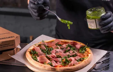 Chef in black gloves adds green pesto sauce to a fresh pizza