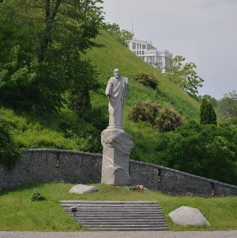 Askoldova Mohyla in Kyiv - majestic stone statue of Apostle Andrew standing on a rock pedestal against the steep lush green slope of the hill